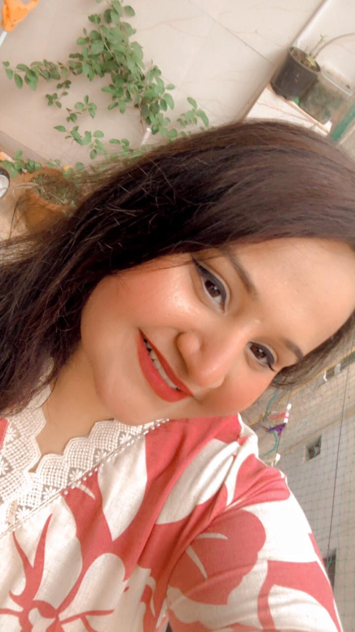 "A woman with long dark hair, wearing a red and white patterned top, smiles brightly at the camera with plants in the background."